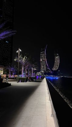 an empty walkway next to the water with palm trees and buildings in the background at night