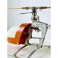 an orange and white helicopter is sitting on the ground next to a wall with a wooden stick sticking out of it's back end