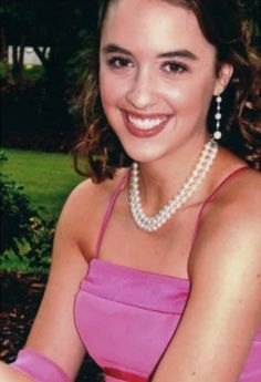 a woman wearing a pink dress and pearl necklace sitting in front of a tree with her hands on her hips