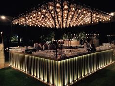 an outdoor bar is lit up at night with lots of lights on the ceiling and people sitting around it