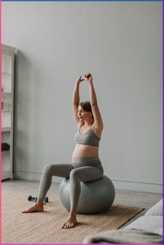 a pregnant woman sitting on an exercise ball in her living room doing yoga with her hands up