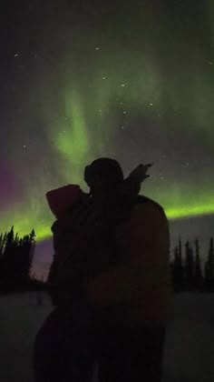 two people are standing in the snow with an aurora light behind them and trees on the other side