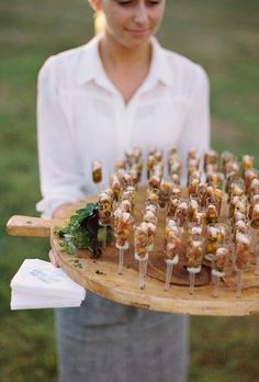 a woman holding a platter full of food on top of a wooden tray in the grass