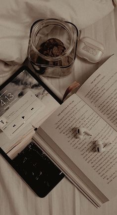 an open book sitting on top of a bed next to ear buds and remotes