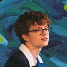 a young man wearing glasses standing in front of a blue and green painting with swirls on it