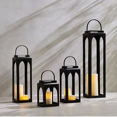 three black lanterns with lit candles in front of a white wall and shadows on the floor