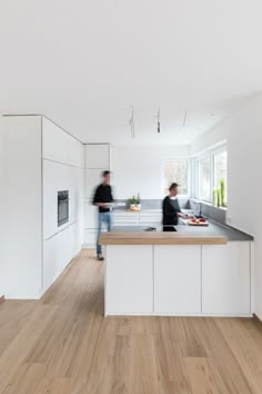 two people standing in a kitchen with white cabinets and wood flooring on the walls