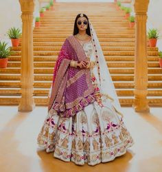 a woman in a white and pink lehenga stands on steps with her hands behind her back