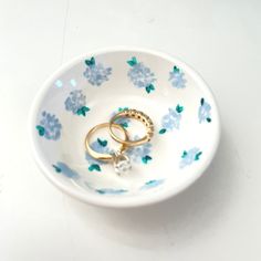 two gold wedding rings in a white bowl with blue flowers on the rim and green leaves