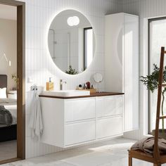 a white bathroom with a large round mirror on the wall and wooden chair in front of it