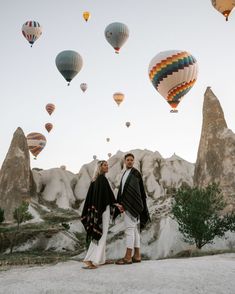 two people standing next to each other in front of hot air balloons flying above them