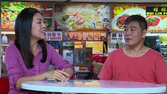 a man and woman sitting at a table in front of a food stand talking to each other