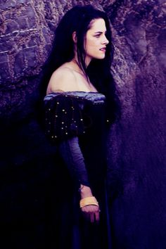 a woman with long black hair standing in front of a rock wall wearing a dress