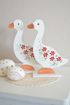 two white ducks are standing next to some eggs on a table with a wall in the background