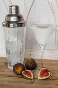 a martini glass next to two figs on a wooden table with a metal shaker