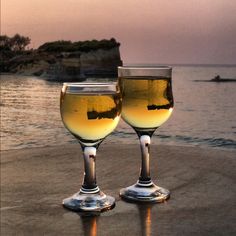 two wine glasses sitting on top of a sandy beach next to the ocean at sunset