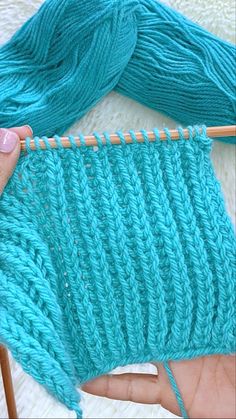 a hand holding a knitting needle over a blue knitted piece of cloth with yarn on it