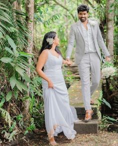 a bride and groom are walking through the woods holding hands while dressed in grey suits