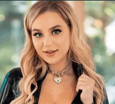 a woman with blonde hair wearing a black leather jacket and silver heart necklace, posing for the camera