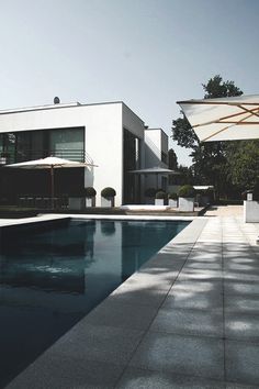 an empty swimming pool in front of a large white house with umbrellas over it