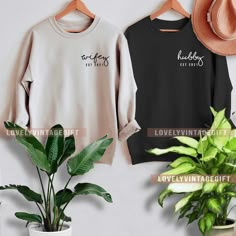 two sweatshirts hanging on a wall next to a potted plant and a hat