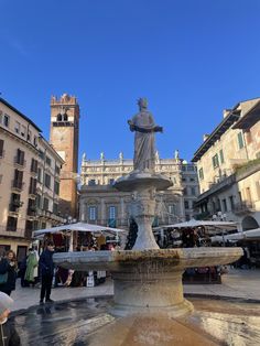 a fountain in the middle of a city square with people walking around and buildings behind it