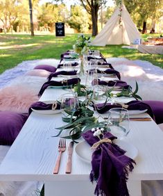 a long table set up with place settings and purple napkins for an outdoor party