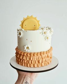 a hand holding a cake decorated with flowers and sun on it's top, in front of a white background