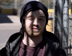 a man with long hair and a beanie looks at the camera while standing in front of a chain link fence