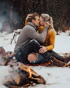 a man and woman sitting next to a campfire in the snow with their arms around each other