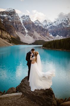 a bride and groom standing on the edge of a mountain lake in front of snow capped mountains