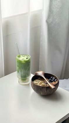 a bowl of cereal and a glass of green juice sit on a table next to a window