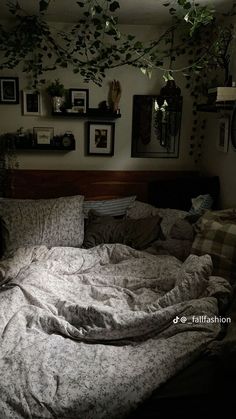 an unmade bed in a bedroom with many pictures on the wall and plants hanging above it