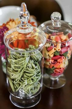 two glass jars filled with different types of pasta and veggies on top of a wooden table