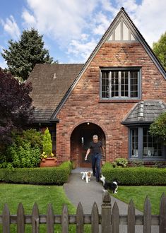 a man walking two dogs in front of a brick house with a picket fence around it