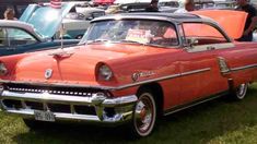 an old red car parked on top of a grass covered field next to other cars