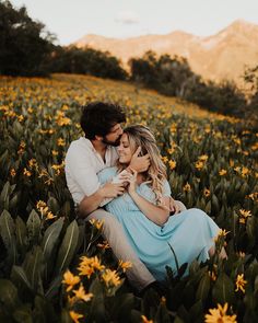 a man and woman are sitting in the middle of a field of flowers with mountains in the background