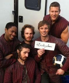 four men posing for a photo in front of a trailer holding a sign that says please to be here