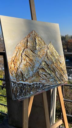 an easel with a painting on it in front of a fence and mountains behind it