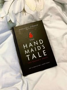 a book sitting on top of a bed next to a white flowered sheet with the title hand maid's tale written in red