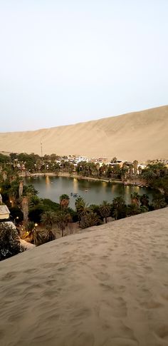 a lake surrounded by sand dunes and palm trees in the distance with buildings on each side