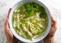 two hands holding a bowl of chicken and celery soup on a marble table