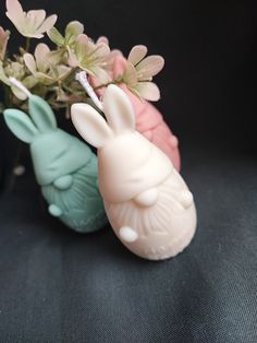 two small plastic rabbits sitting next to each other on a black surface with flowers in the background