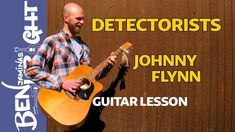 a man holding an acoustic guitar in front of a wooden wall with the words, detectorists johnny flyn guitar lesson