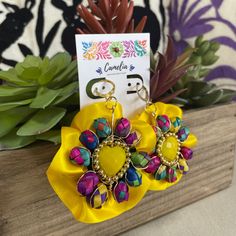 a pair of yellow and pink flower earrings on top of a wooden box next to succulents