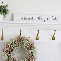 a wreath is hanging on the wall next to two hooks and a sign that says come in and stay awhile