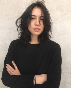 a woman with her arms crossed standing in front of a white wall wearing a black sweater