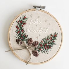 a cross stitch christmas wreath with pine cones and red berries is hanging from a hoop