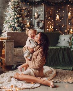 a woman holding a baby in her arms while sitting on the floor next to a christmas tree