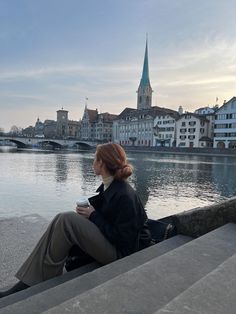 a woman is sitting on the edge of some steps looking at the water and buildings